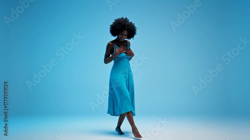 A person with afro hair, wearing a teal dress, dancing barefoot against a blue backdrop