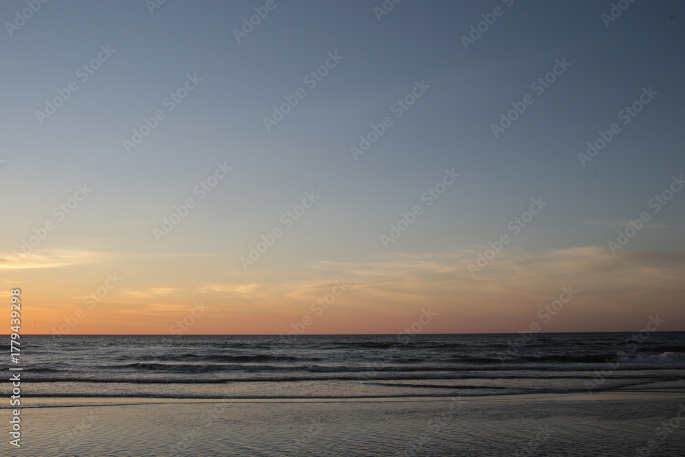 Fototapeta premium A minimalist view of a vast, clear blue-to-orange twilight sky over the dark, calm ocean and beach.