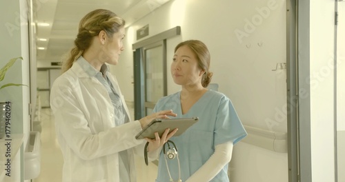 Pointing doctor in lab coat guiding nurse viewing digital tablet in hospital hall, with stethoscope