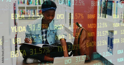 Writing student referencing laptop, notebook and pen in library aisle, with financial data overlay