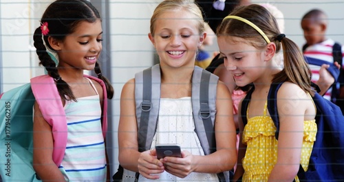 Smiling schoolgirls in dresses gathering around smartphone in school corridor, with three backpacks
