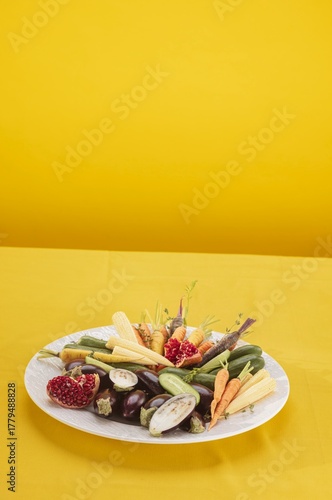 A White plate holds a colourful arrangement of fresh raw vegetables, including mini eggplants, baby carrots, cucumbers, and pomegranates, all on a bright yellow background