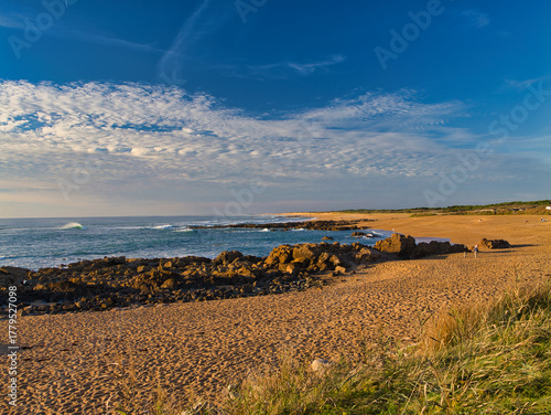 Warm sunset light bathes a wide sandy shore and rocky outcrops at Les Sables-dOlonne on the Vendee coast, France, with gentle waves and patterned clouds.