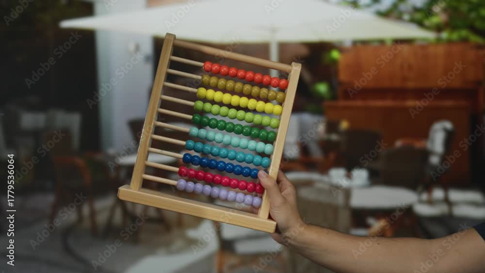 Man holding colorful abacus on outdoor restaurant terrace with blurred background showcasing tables and chairs, evoking a playful educational scene in a city setting.