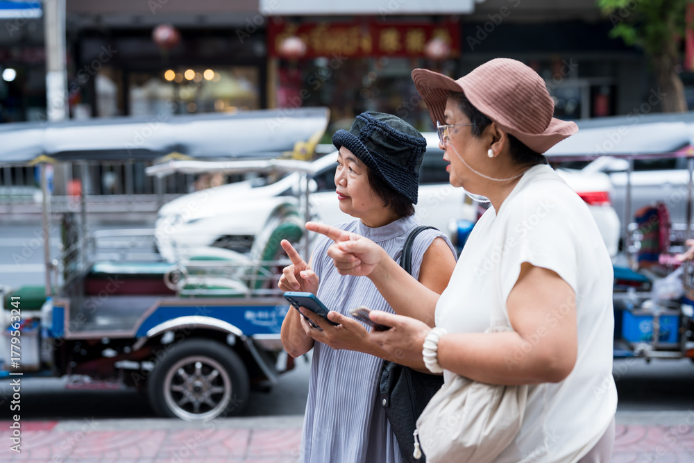 Naklejka premium Two senior asian women travelers holding smartphone and pointing while discussing direction beside tuk tuk showing friendship and city travel lifestyle happiness and active retirement tourism
