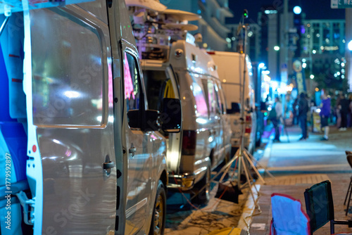 Multiple television news vans line a downtown street with satellite dishes and lights. Crews prepare mobile broadcast units for live transmission and media coverage.