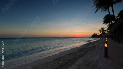 Sunset view on Eagle Beach resort shoreline with calm waves and palm trees creating tropical evening background. Aruba.