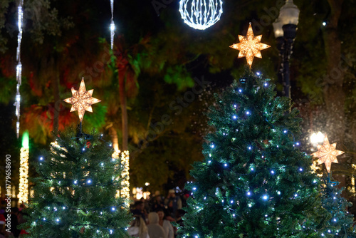 Two evergreen trees decorated with white LEDs and glowing star toppers shine in a park. Festive holiday scene with snowfall effects and bright bokeh.