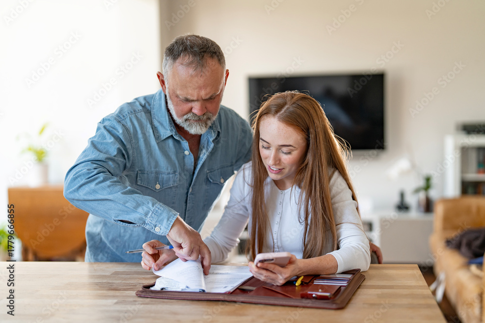 Fototapeta premium Elderly man reviews documents with younger woman in bright living room while she focuses on her phone