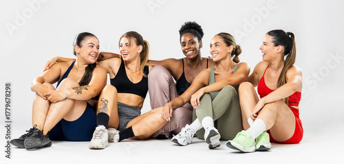 A group of 5 girls of different ethnicities are posing in colorful sportswear against a white background.The girls are sitting happily hugging each other.Concept of women in sports.