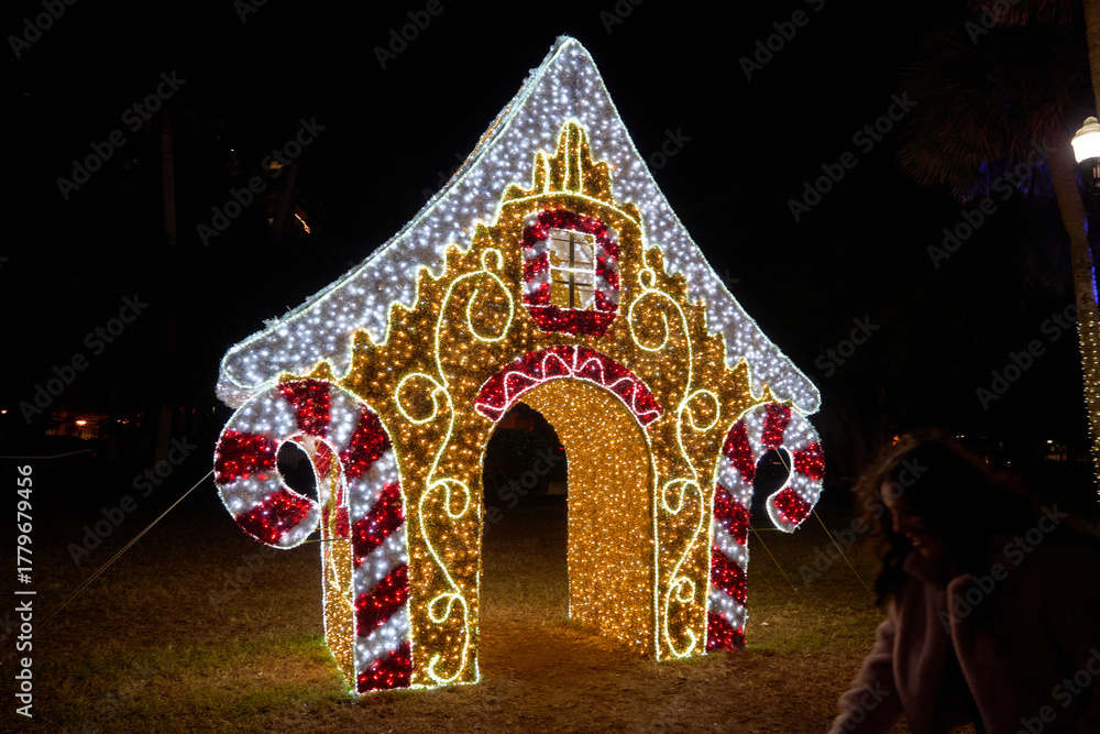 Naklejka premium A large gingerbread house arch made of LED lights glows at night. Festive candy cane columns and bright decoration welcome families in a winter park.
