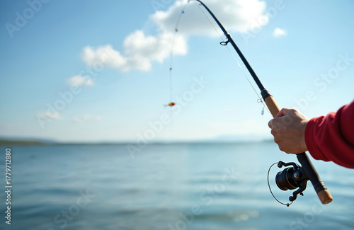 Fototapeta Naklejka Na Ścianę i Meble -  Person in red shirt holds a fishing rod, casting a lure over calm water. Bright blue sky with clouds above, distant land. Man enjoys outdoor hobby, angling sport near lake or river shore.