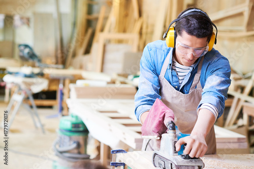 Young adult Caucasian man wearing protective goggles and earmuffs using electric planer on wooden board in carpentry workshop, focusing on precise woodworking task