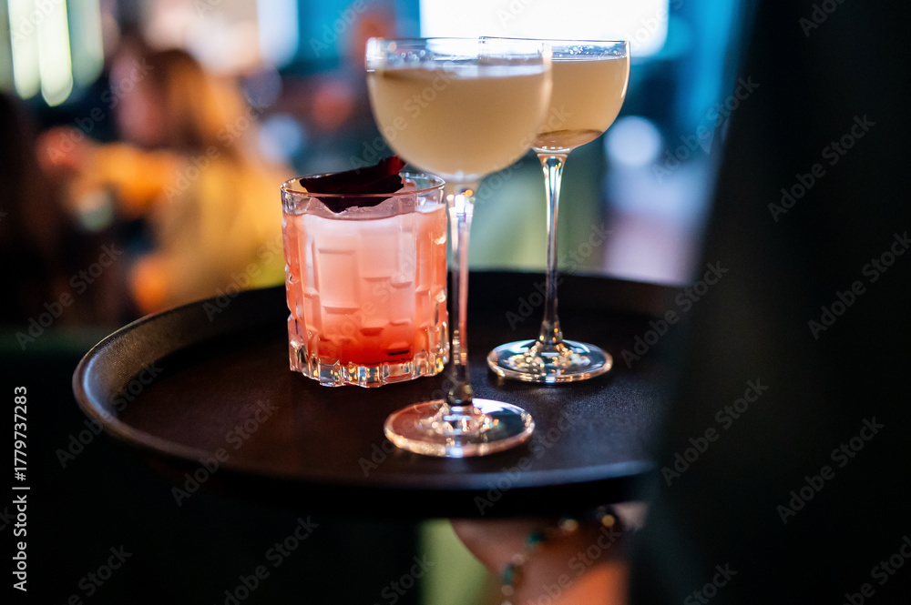 Fototapeta premium A server holds a black tray with three cocktails. One is a pink drink in a rocks glass, garnished with a dark leaf. The other two are pale yellow in classic coupe glasses