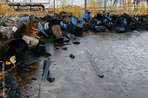 Industrial site with discarded barrels and debris, surrounded by overgrown vegetation, reflecting environmental neglect and pollution in a desolate landscape with water accumulation
