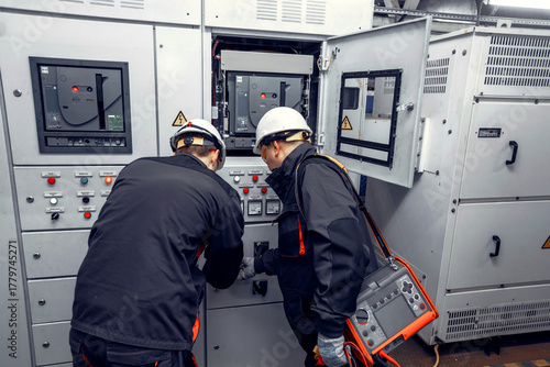 Two technicians, wearing safety helmets and work attire, are inspecting electrical control panels in a modern industrial facility, showcasing teamwork and technical expertise in electrical maintenance