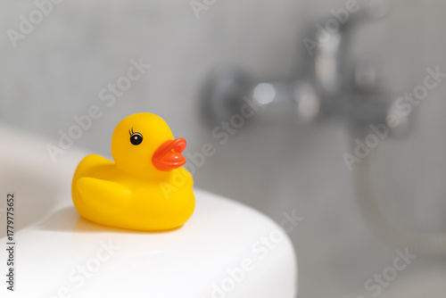 Yellow rubber duck on the corner of a white bathroom sink, photographed from three-quarter angle, blurred faucet and blue-gray tiles in background, close-up concept of hygiene and childhood