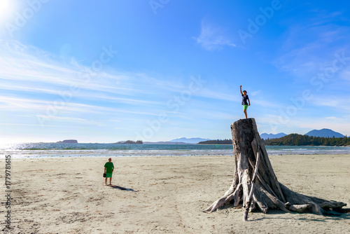 Teenager boy climbed the tall driftwood stump in the sandy beach, dad is watching him. Sunny day at the pacific coast, Tofino, Vancouver island, BC, Canada.