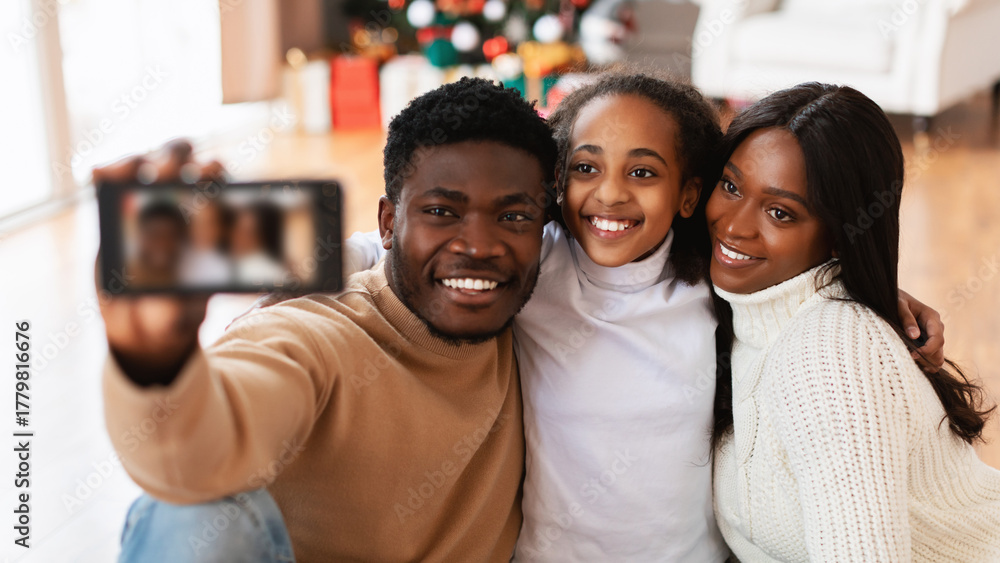 Obraz premium A happy black family enjoys quality time together, celebrating Christmas at home. They take a cheerful selfie with their excited daughter hugging her parents, all smiling and looking at the camera.