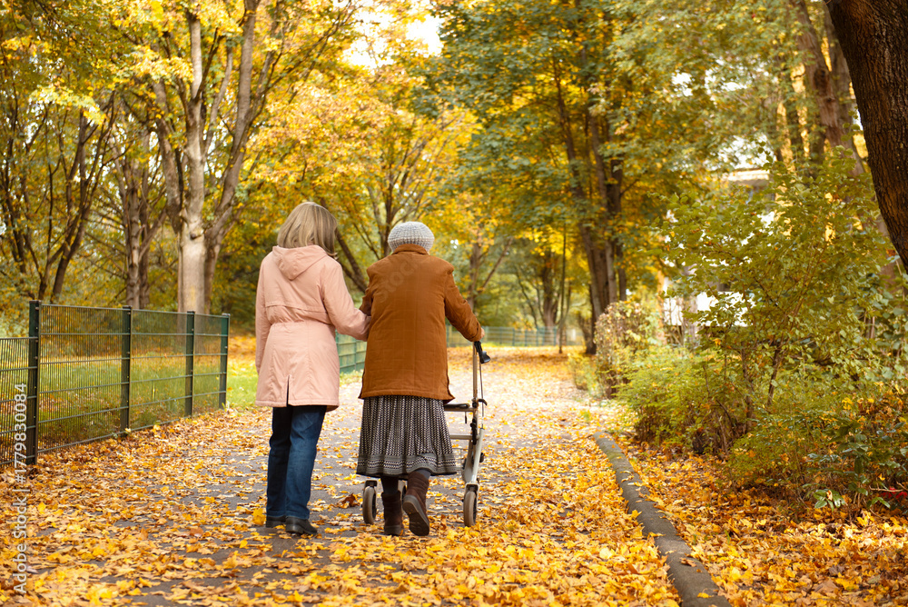 Fototapeta premium Woman and her elderly mother with walker taking a walk in the park covered in autumn leaves, spending family time together
