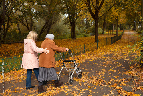 Woman and her elderly mother with walker taking a walk in the park covered in autumn leaves, spending family time together