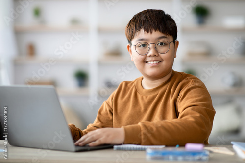 Chubby smart boy is focused on his homework at a table in a well-lit room. He types on his laptop while surrounded by school supplies, engaging in online learning.