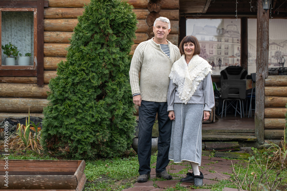 Fototapeta premium Village couple pensioners posing near a log bathhouse.