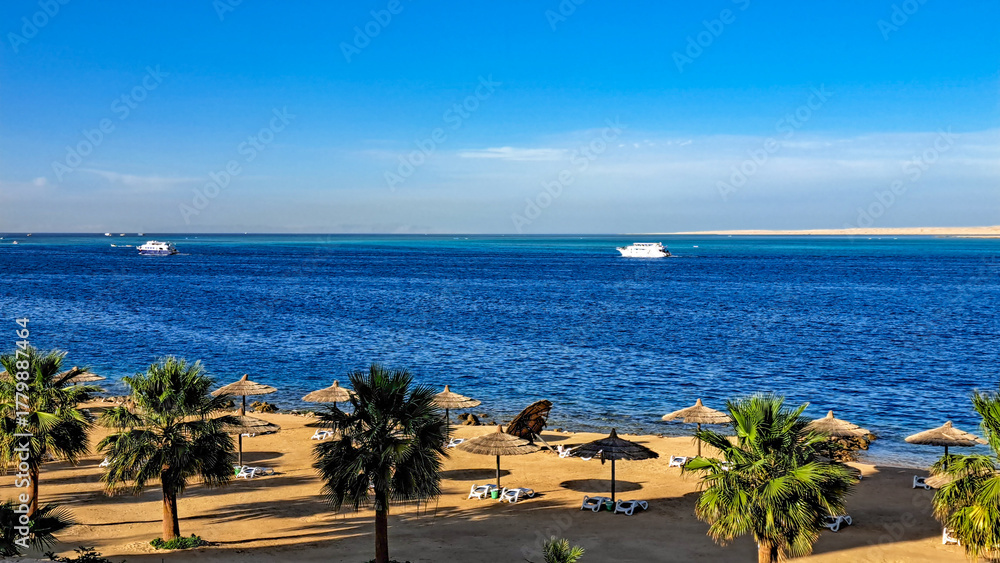 Obraz premium Scenic beach view with palm trees and umbrellas overlooking tranquil blue waters, boats in the distance, creating a serene coastal atmosphere for relaxation and leisure. Hurghada, Egypt, Red Sea