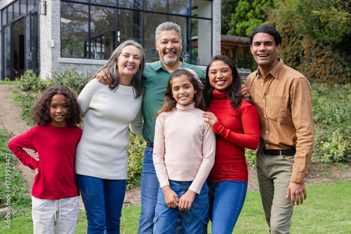 Diverse extended family standing close together on front lawn of suburban house smiling at camera