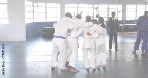 Cheering martial arts team gathering in training hall, lifting gold trophy and wearing white gis
