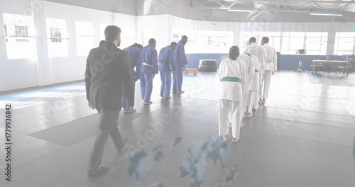 Walking instructor in dark suit approaching bowing martial artists on dojo mats, with gis and belts