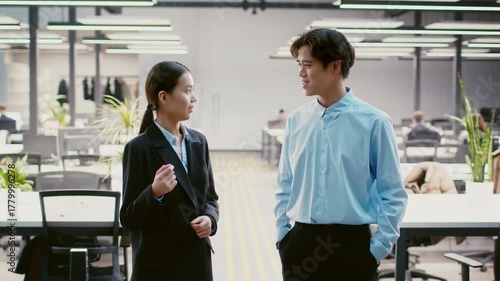 Two young Asian professionals converse in a contemporary office environment filled with plants and natural light. Their attire suggests a formal work setting, indicative of a collaborative atmosphere.