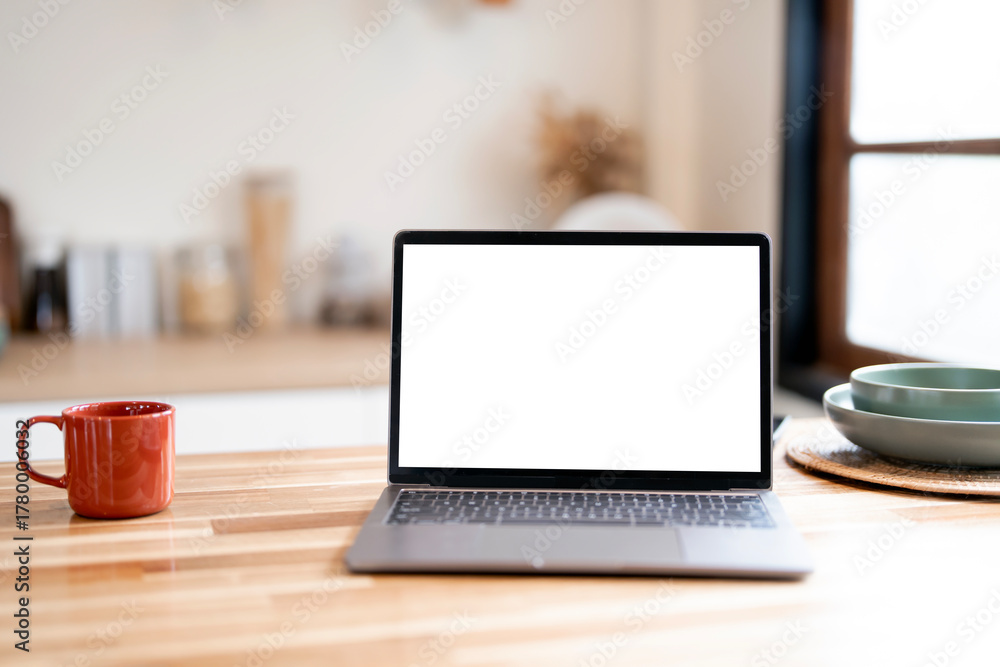 Fototapeta premium Laptop with a blank white screen placed on a wooden table next to a coffee mug and breakfast bowls in a home kitchen setting.