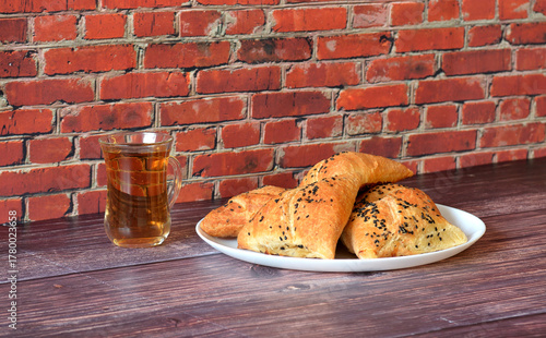 Large plate with several meat pies samsa decorated with caraway seeds and a cup of hot tea on a wooden table.