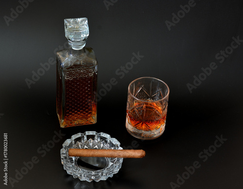A faceted glass and a crystal decanter with old whiskey and a glass ashtray with a Cuban cigar on a black background.
