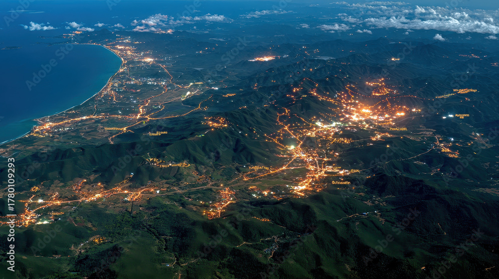 Fototapeta premium Coastal cityscape at night illuminated by streetlights with mountain ranges surrounding urban areas and glowing lights reflecting on calm ocean shore under partly cloudy sky.