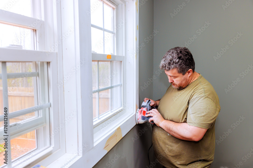 Fototapeta premium Handyman is carefully sanding wood trim next to window in well lit room on task, preparing for painting finishing in home improvement