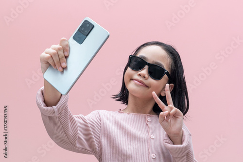 Confident Asian young girl wearing sunglasses taking a selfie with a smartphone. Generation Alpha, technology, digital lifestyle, and social media culture.