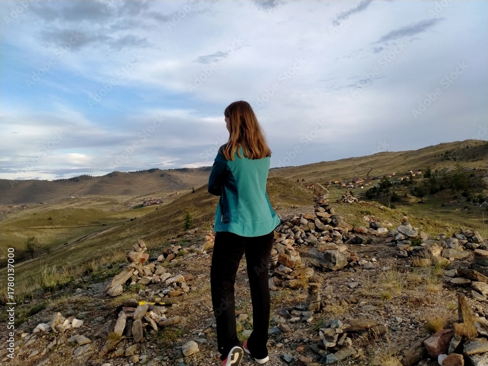 Naklejka premium A woman on the crest of a hill admires the wide steppe expanses in the soft evening light. Stone slopes and a village in the distance, a feeling of freedom.