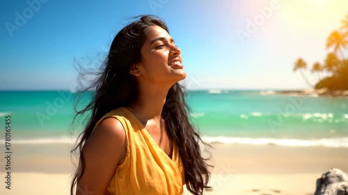 Beautiful young Indian woman wearing a flowing yellow dress, smiles joyfully on tropical beach. She looks up at the bright sun. Evokes feelings of happiness and enjoying summer vacation