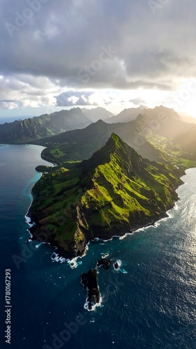 Aerial view of a lush, verdant mountain range meeting the deep blue ocean, with sunlight breaking through dramatic clouds