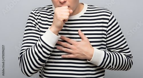 Close-up of adult coughing with hand on chest in striped shirt, Person covering mouth while experiencing chest discomfort, Studio shot showing cough or breathing difficulty concept, Health and illness