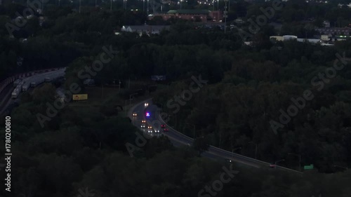 Police car with flashing lights on highway overtaking cars in suburbia. Aerial wide shot. Dusk scene in the evening. Forest trees in fall season. America, Virginia.
