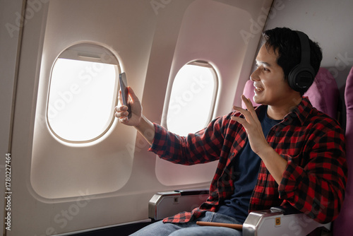 Young Asian man sitting on airplane seat taking a selfie with his smartphone near the window, smiling and wearing wireless headphones. Travel lifestyle and capturing memories during flight.
