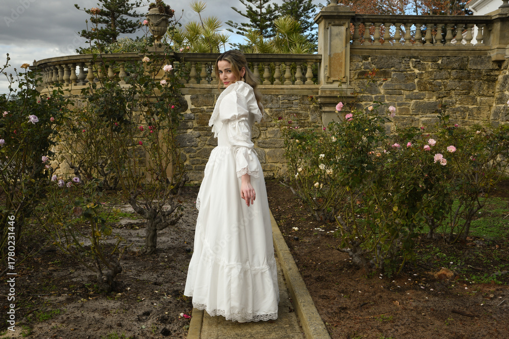 Fototapeta premium portrait of beautiful young blonde model wearing historical white wedding gown. ethereal back view figure, walking away in romantic rose garden of fairytale castle garden background