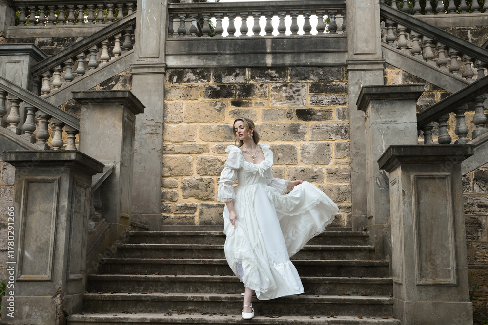 Naklejka premium Full length female figure of blonde model wearing white fantasy wedding gown. walking down stairs on historical staircase of fairytale castle location 