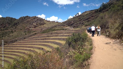 Group of tourists following a guide along a dirt path past vast Inca agricultural terraces and ruins at the Pisac Archaeological Park, Peru