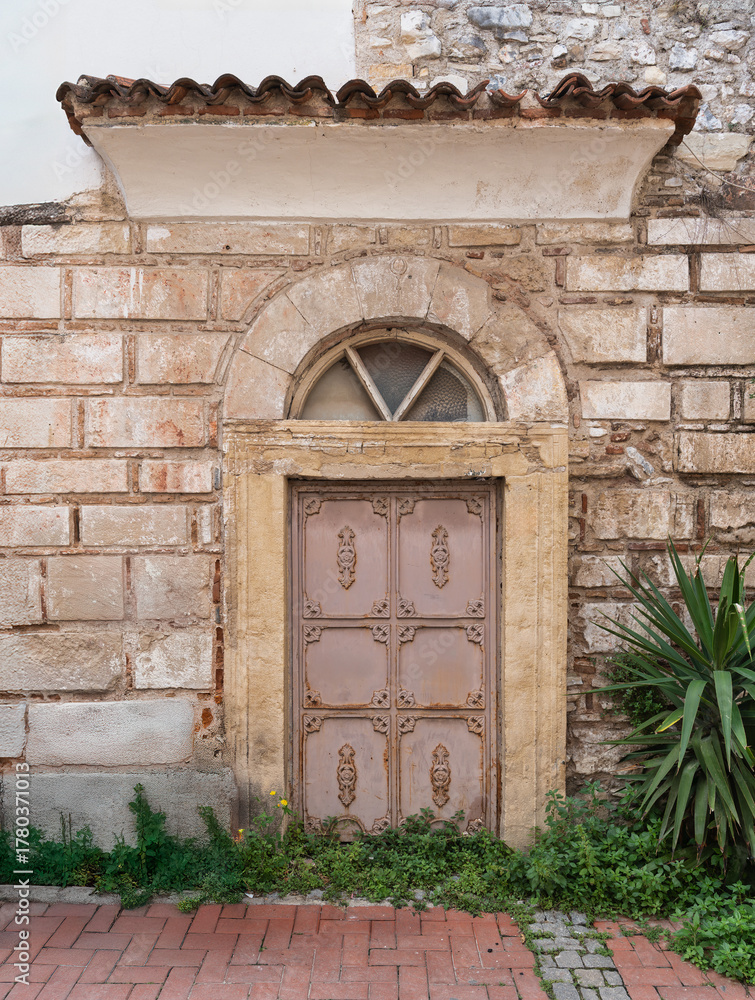 Fototapeta premium Historic ornate metal door with arched window, crescent moon carving, and tiled roof on a stone wall. Green plants, brick pavement