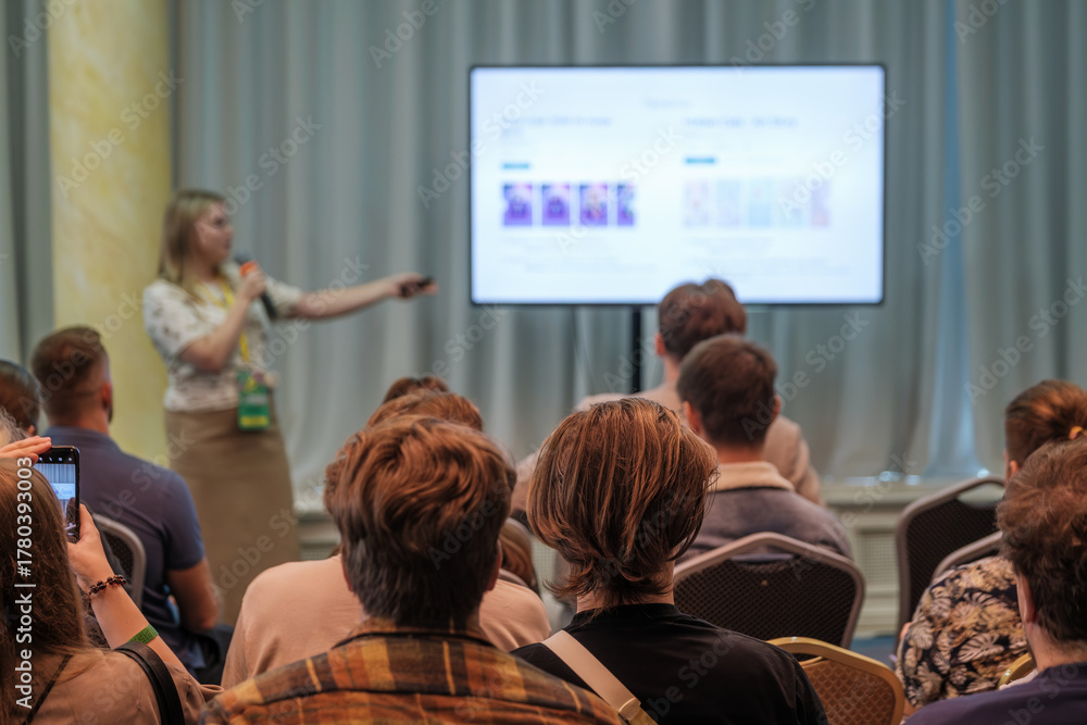 Obraz premium Presenter speaks into microphone while pointing to projected slide as attendees watch. Bright room, conference setting, engaged audience, learning vibe, professional presentation, sharing information.