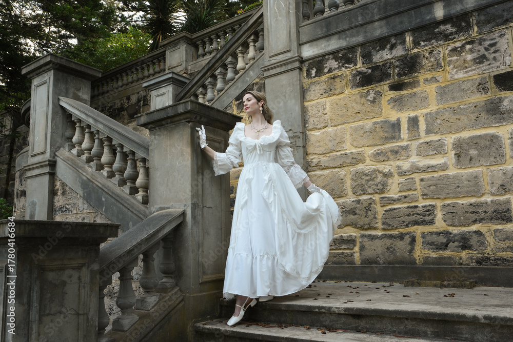 Obraz premium Full length female figure of blonde model wearing white fantasy wedding gown. walking down stairs on historical staircase of fairytale castle location 
